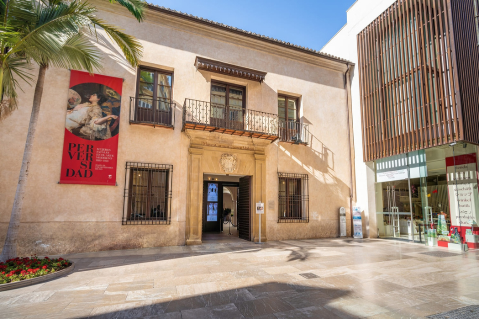 Jasnobeżowa fasada muzeum z ozdobnym wejściem, balkonem, czerwonym banerem wystawy i palmą